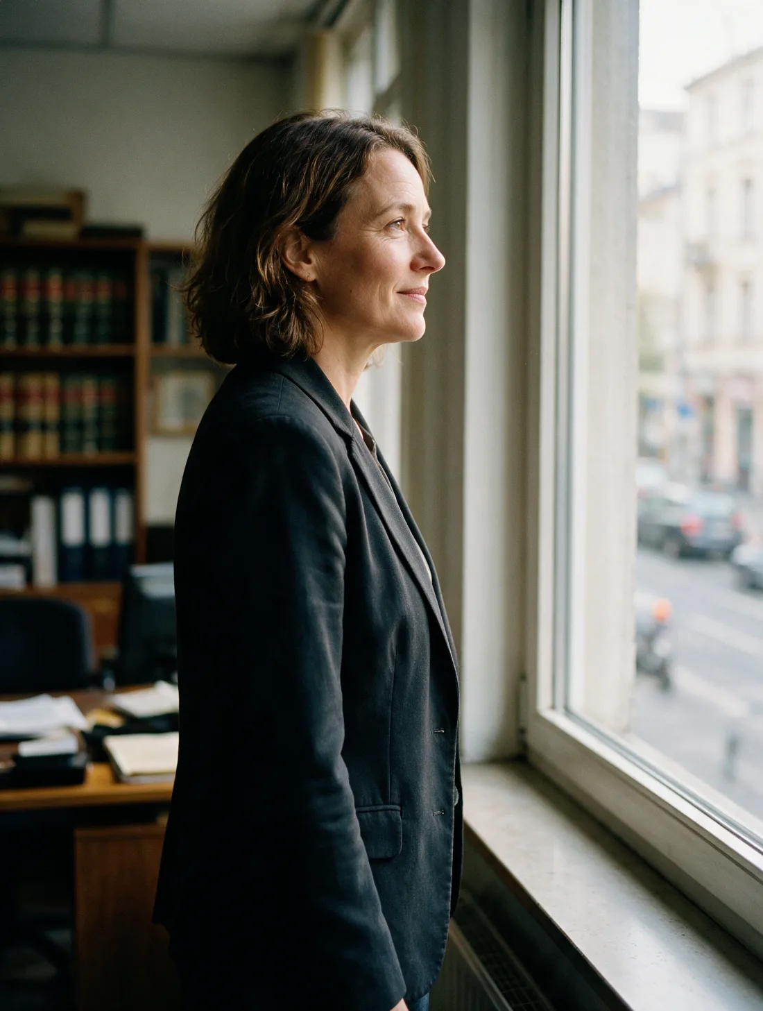 Eine Frau im dunklen Blazer steht im Profil am hohen Bürofenster und schaut auf die Straße.